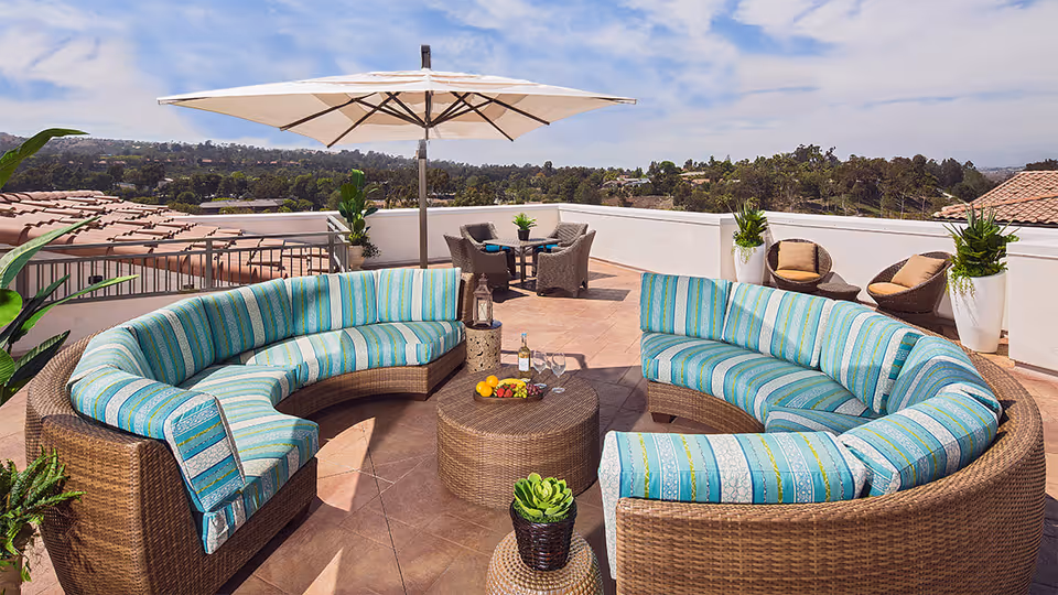 Outdoor patio area with two curved wicker sofas featuring blue and green striped cushions, a round wicker coffee table with a bottle of wine, glasses, and fruit, potted plants, two wicker chairs with cushions, and a large white umbrella. The background shows a scenic view of trees and hills under a partly cloudy sky.