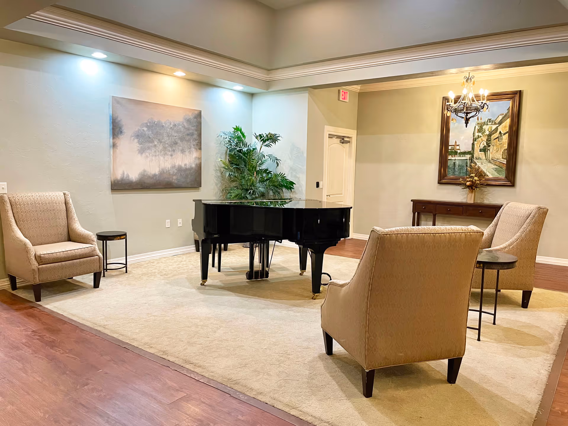 A cozy interior room with a black grand piano in the center on a large beige rug. There are two beige upholstered armchairs facing the piano, each with a small round side table next to it. The walls are light-colored with crown molding, and two framed paintings hang on the walls. A green potted plant is placed near the piano, and a chandelier hangs from the ceiling.
