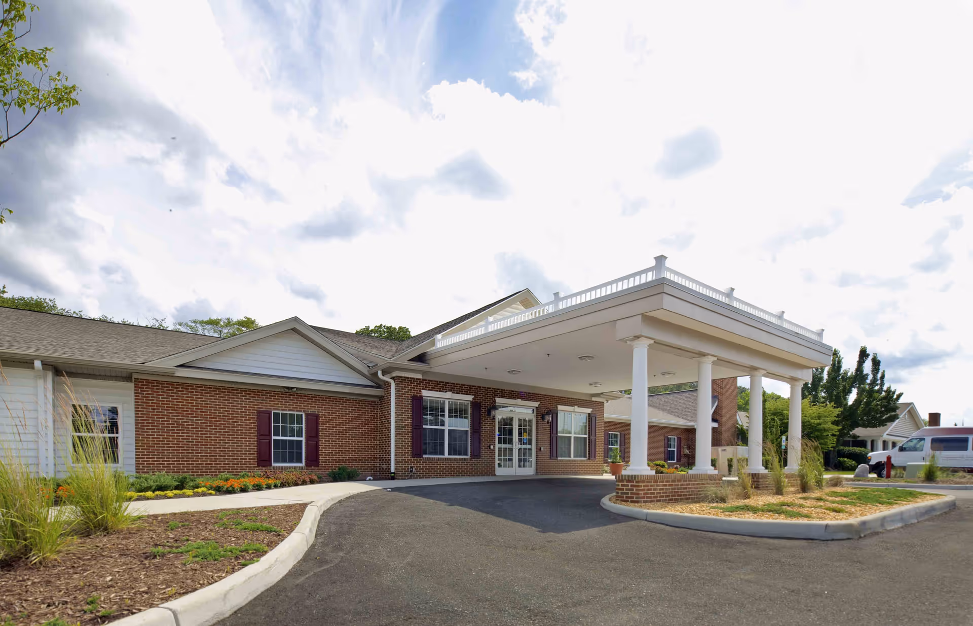Exterior view of Commonwealth Senior Living at Hampton showing a single-story brick building with white columns supporting a covered entrance. The building has multiple windows with maroon shutters and a landscaped area with plants and flowers near the entrance. The sky is partly cloudy.