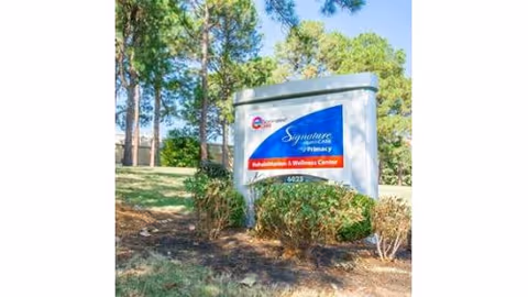 Outdoor view of a sign for Signature HealthCARE of Primacy Rehabilitation & Wellness Center surrounded by bushes and trees with a clear blue sky in the background.