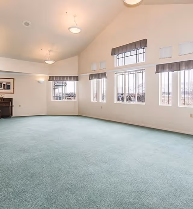 A spacious, empty room with light green carpet, cream-colored walls, and multiple large windows with gray valances allowing natural light to fill the space. The ceiling is vaulted with two hanging light fixtures, and there is a framed picture and a small piece of furniture against one wall.