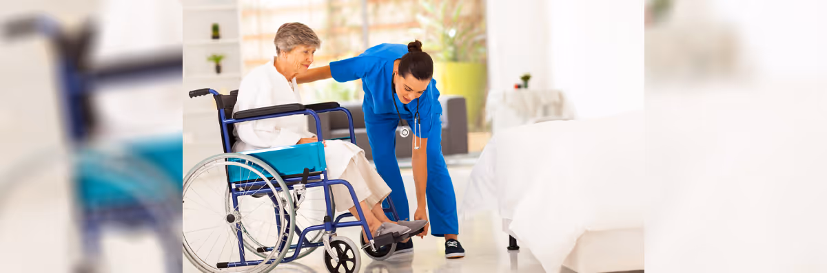 A healthcare worker in blue scrubs is assisting an elderly woman in a wheelchair by adjusting her shoe in a bright, clean bedroom setting.
