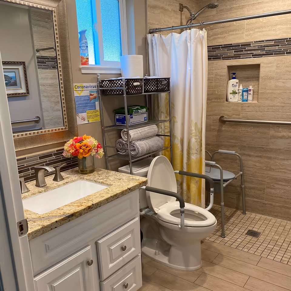 Well-equipped accessible bathroom featuring a vanity sink, toilet with arm rails, shower with grab bar and chair, and shelving with towels and supplies.