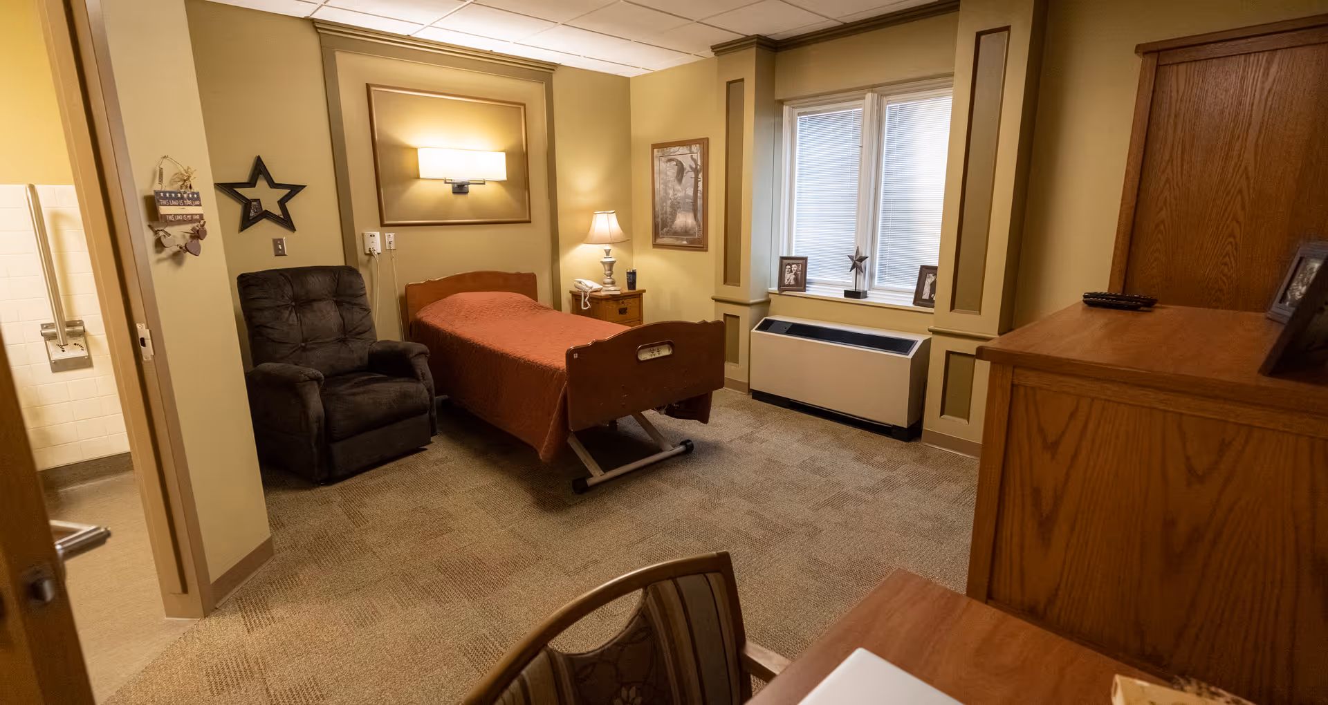 A single bedroom in a senior living facility with a hospital-style bed covered with a brown blanket, a dark recliner chair, a wooden nightstand with a lamp and phone, a window with blinds, framed pictures on the windowsill, and a wooden dresser. The room has beige walls and carpeted floor. A bathroom is partially visible through an open door on the left.