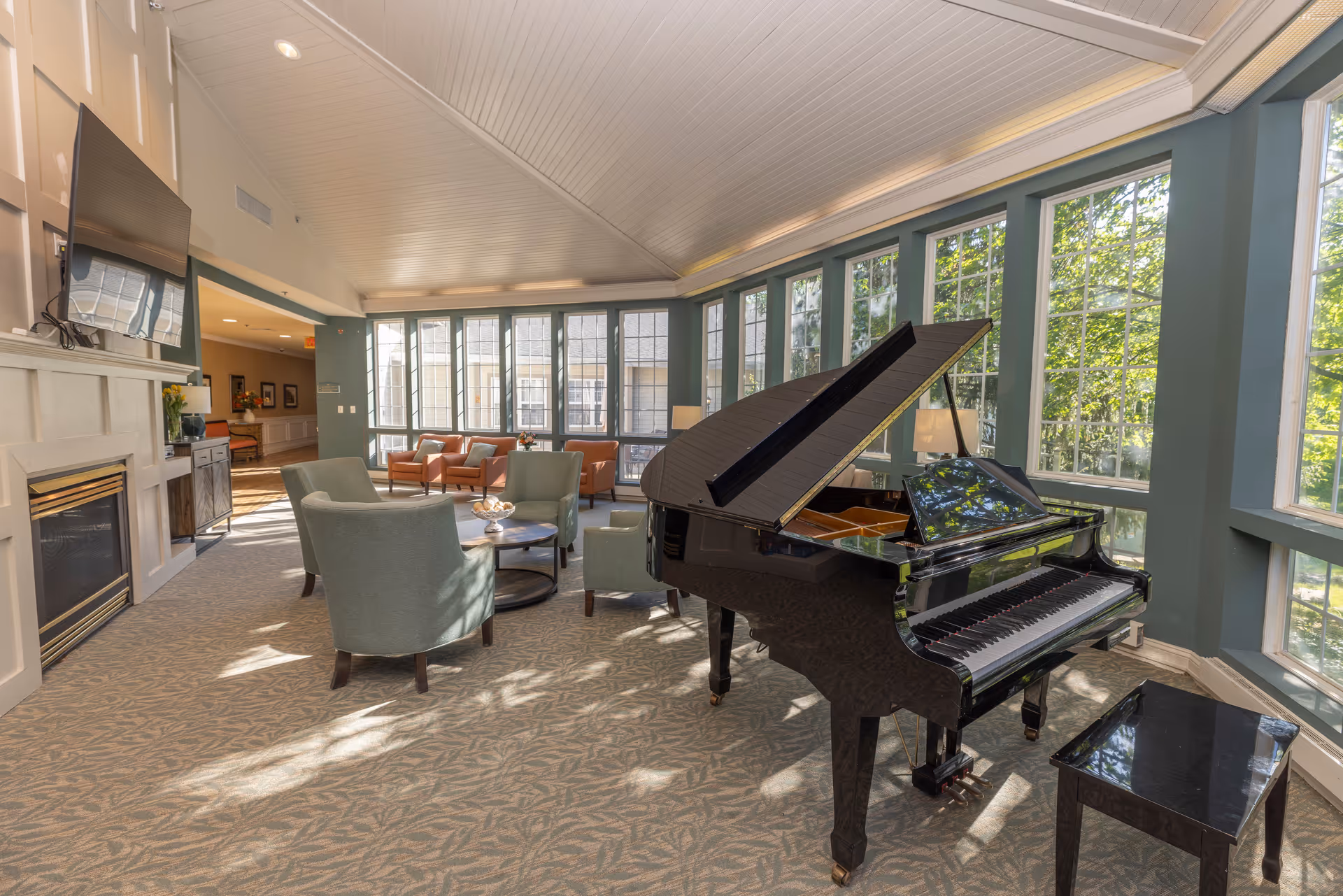 A bright and spacious living room area with large windows allowing natural light to fill the space. The room features a black grand piano with a matching bench, several comfortable armchairs arranged around a small round table, a fireplace with a mounted flat-screen TV above it, and additional seating near the windows. The ceiling is vaulted with white paneling, and the carpet has a subtle leaf pattern.