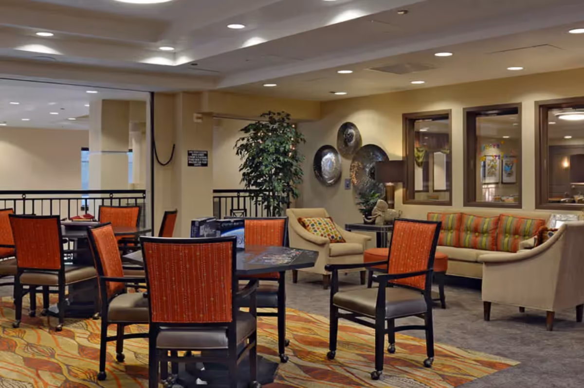 Well-lit communal lounge/dining area with tables surrounded by orange-upholstered chairs, sofas, and decorative wall art.