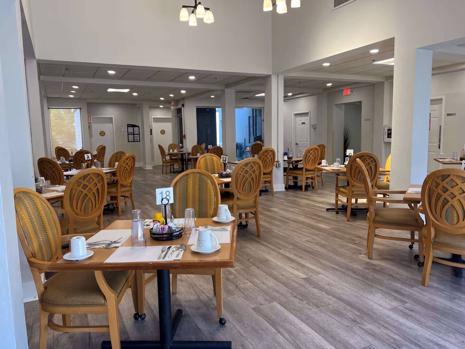 Spacious dining room in a senior living facility with multiple set wooden tables and chairs on wood-look flooring.