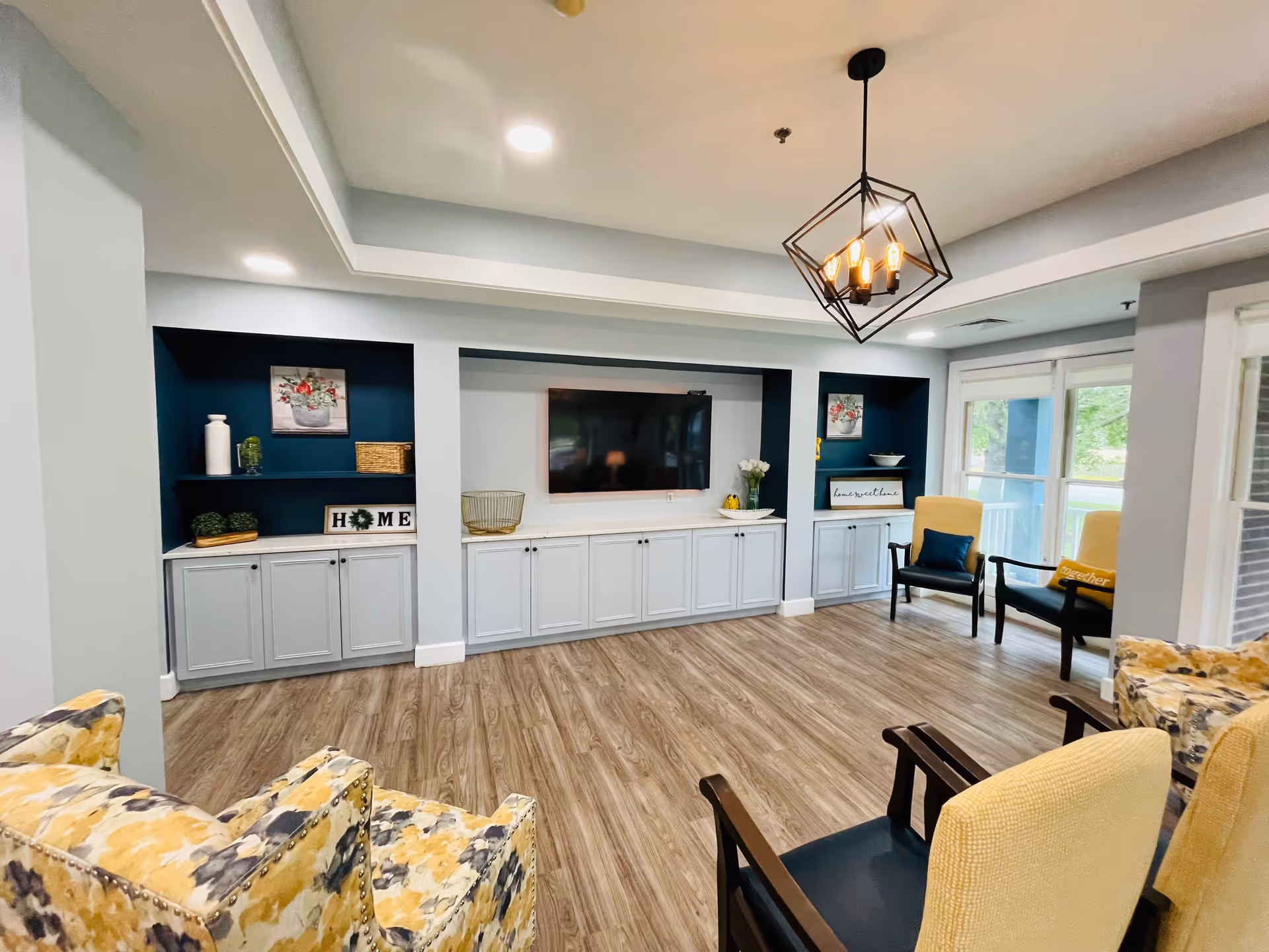 A bright and modern living room with wood flooring, a mounted flat-screen TV on a wall with built-in white cabinets and dark blue shelves. The shelves are decorated with vases, plants, and signs that say 'HOME' and 'home sweet home'. There are several upholstered chairs in yellow and floral patterns arranged around the room, and large windows letting in natural light. A geometric pendant light hangs from the ceiling.