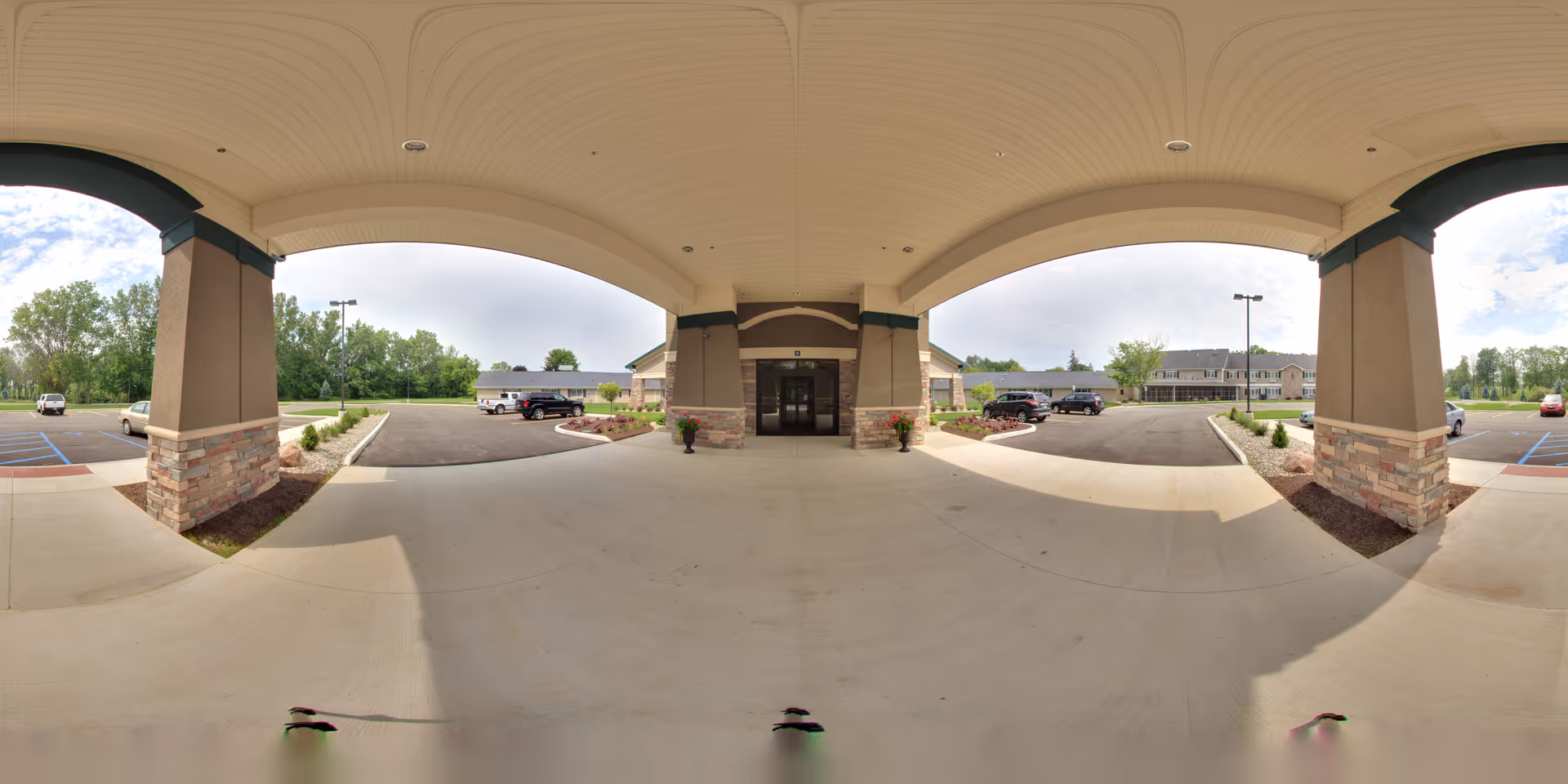 Covered entrance area of a senior living facility with a wide concrete driveway and parking spaces on both sides. The building entrance is centered with double glass doors, flanked by stone and beige pillars. There are landscaped areas with small shrubs and flowers near the entrance. Several cars are parked in the lot, and trees and greenery are visible in the background under a partly cloudy sky.