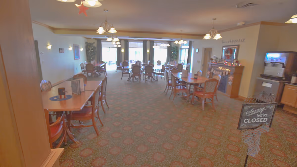 A dining room in a senior living facility with multiple wooden tables and chairs arranged neatly. The room is well-lit with hanging light fixtures and has large windows at the far end letting in natural light. There is a fireplace decorated with lights and stars on the right side, and a sign near the entrance that reads 'Sorry We're Closed'.