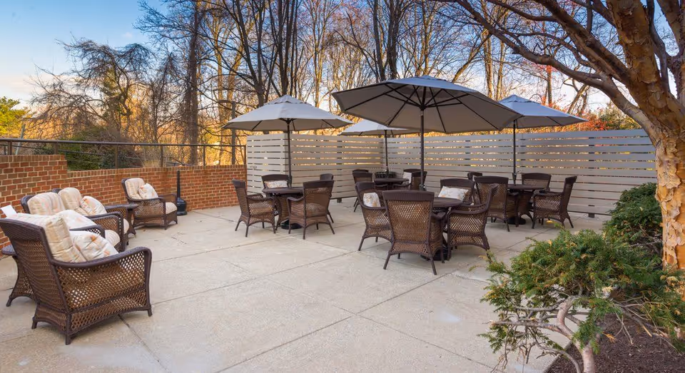 Outdoor patio area with several wicker chairs and tables, each table shaded by large umbrellas. The patio is surrounded by a brick wall and a wooden privacy fence, with trees and shrubs visible around the perimeter.