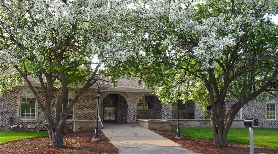 Brick senior living facility front entrance framed by flowering trees along a walkway.