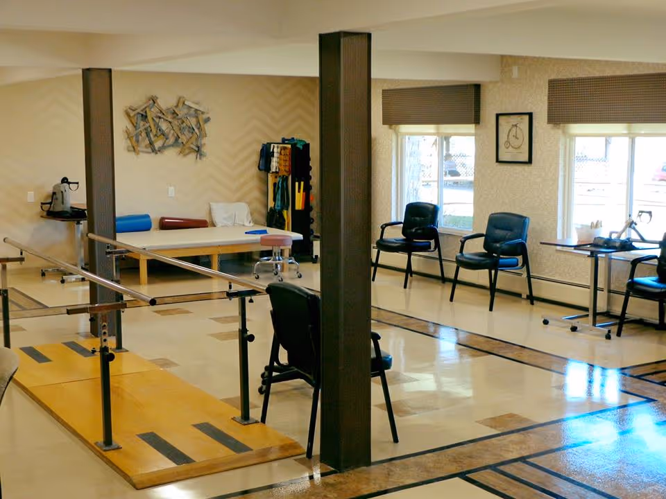 Rehabilitation therapy room with parallel walking bars, chairs, and a treatment table in a senior living facility.