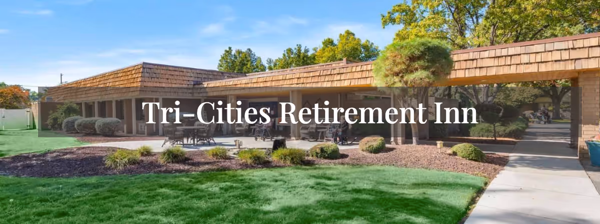 Outdoor view of Tri-Cities Retirement Inn showing a single-story building with a shingled roof, surrounded by green grass, landscaped bushes, and trees under a clear blue sky. There are outdoor tables and chairs on a patio area.