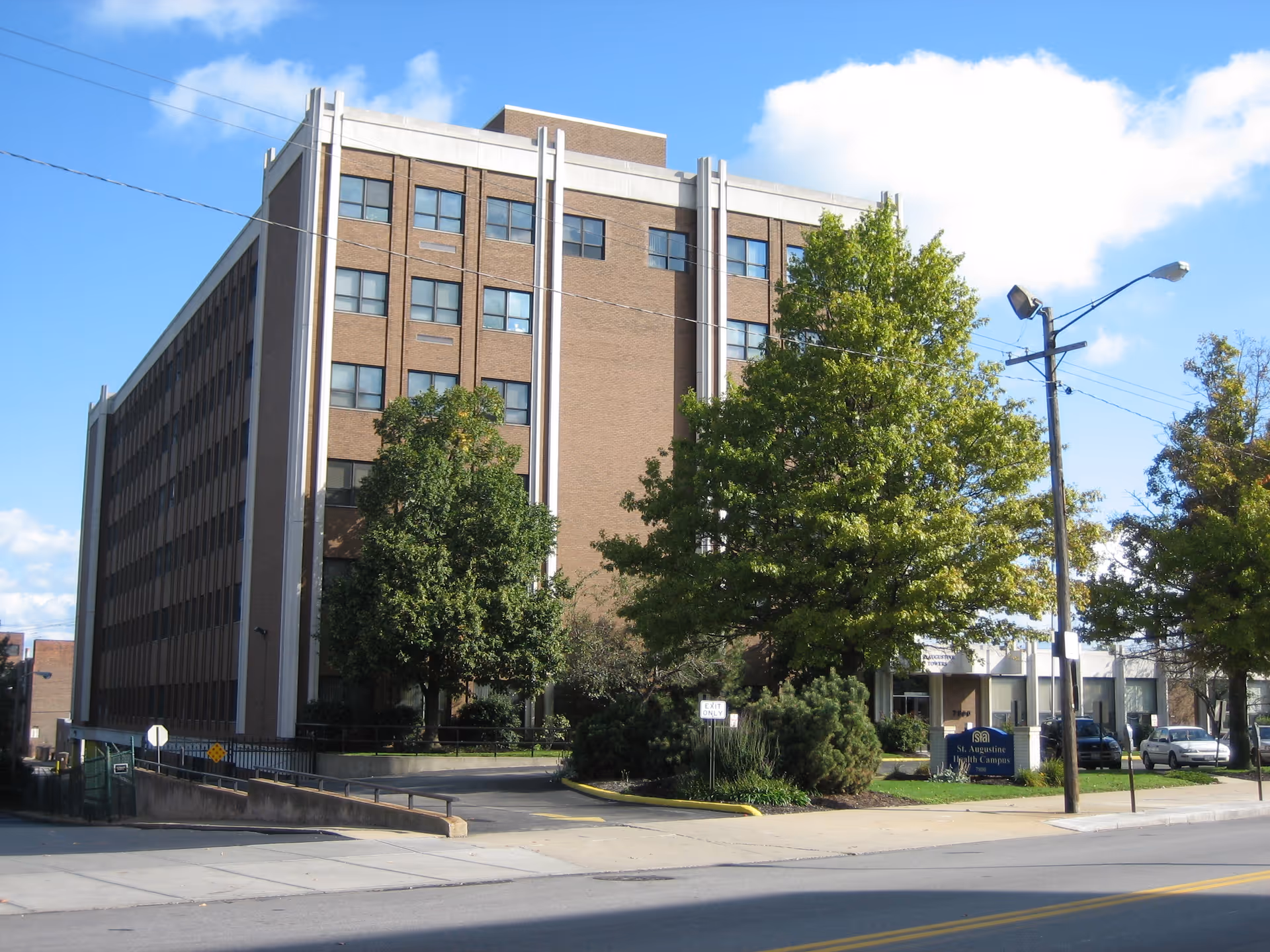 Exterior view of a multi-story brick building with several windows, surrounded by trees and a parking area. A sign in front reads 'St. Augustine South Campus'. The sky is clear with a few clouds.