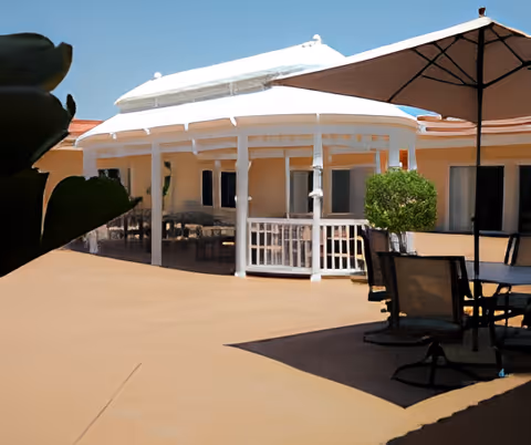 Outdoor patio area with a white gazebo structure, patio tables and chairs, and a large umbrella providing shade. The background shows part of a building with windows and a clear blue sky.