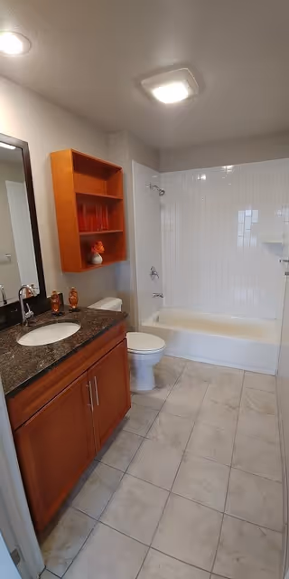A clean bathroom featuring a white bathtub with a tiled shower surround, a toilet, and a wooden vanity with a dark granite countertop and a sink. Above the toilet is a wooden wall-mounted cabinet with decorative items. The floor is tiled in a light color, and the ceiling has two light fixtures.