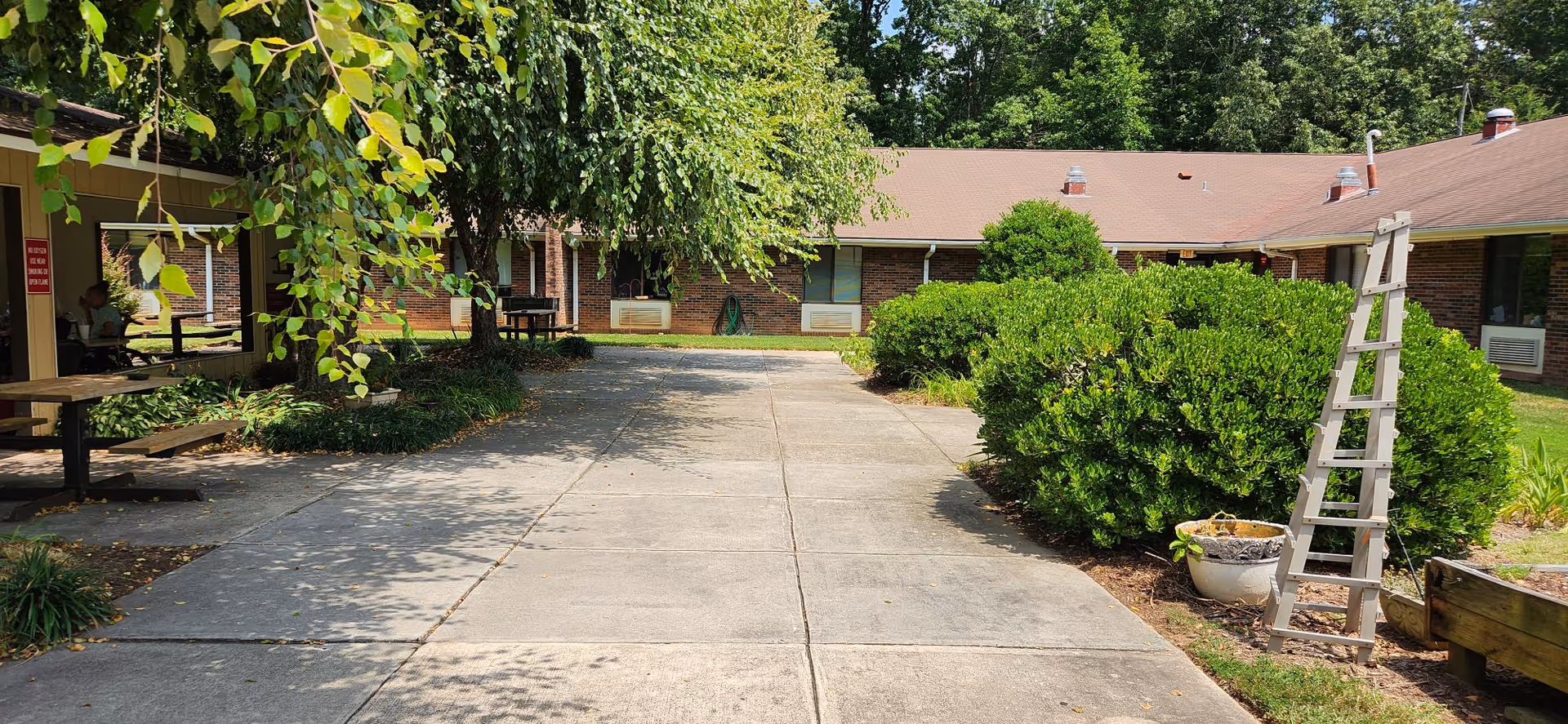 Outdoor courtyard area with concrete walkway, green bushes, trees, and picnic tables. The surrounding building has a brick exterior with windows and a brown roof. There is a wooden ladder and a potted plant on the right side.