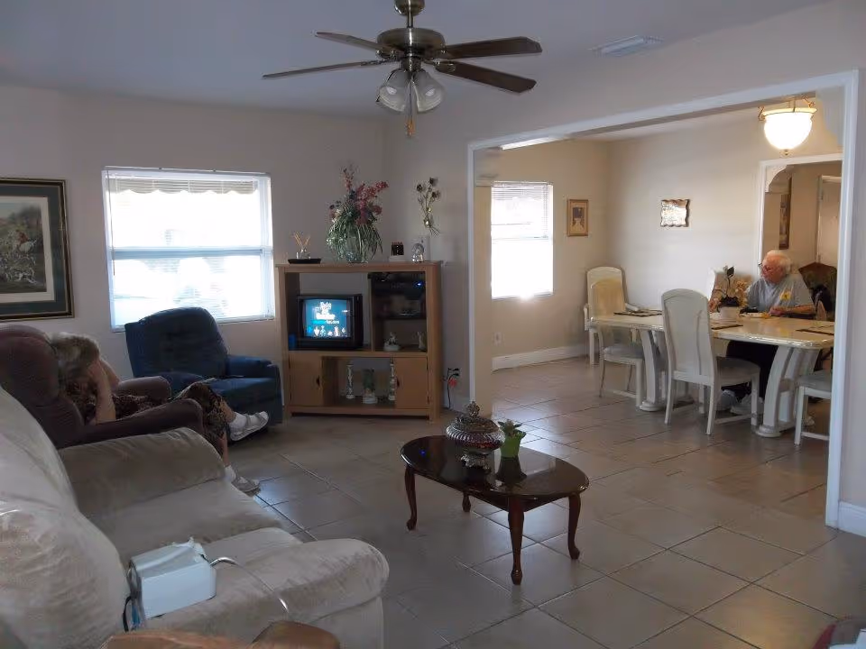 A living room and dining area in a senior living facility. The living room has a beige couch, a blue recliner, a wooden TV stand with a small TV, and a ceiling fan with lights. There is a coffee table with decorative items. The dining area has a white dining table with chairs, where an elderly person is seated and eating. The walls are light-colored with framed pictures, and the floor is tiled.