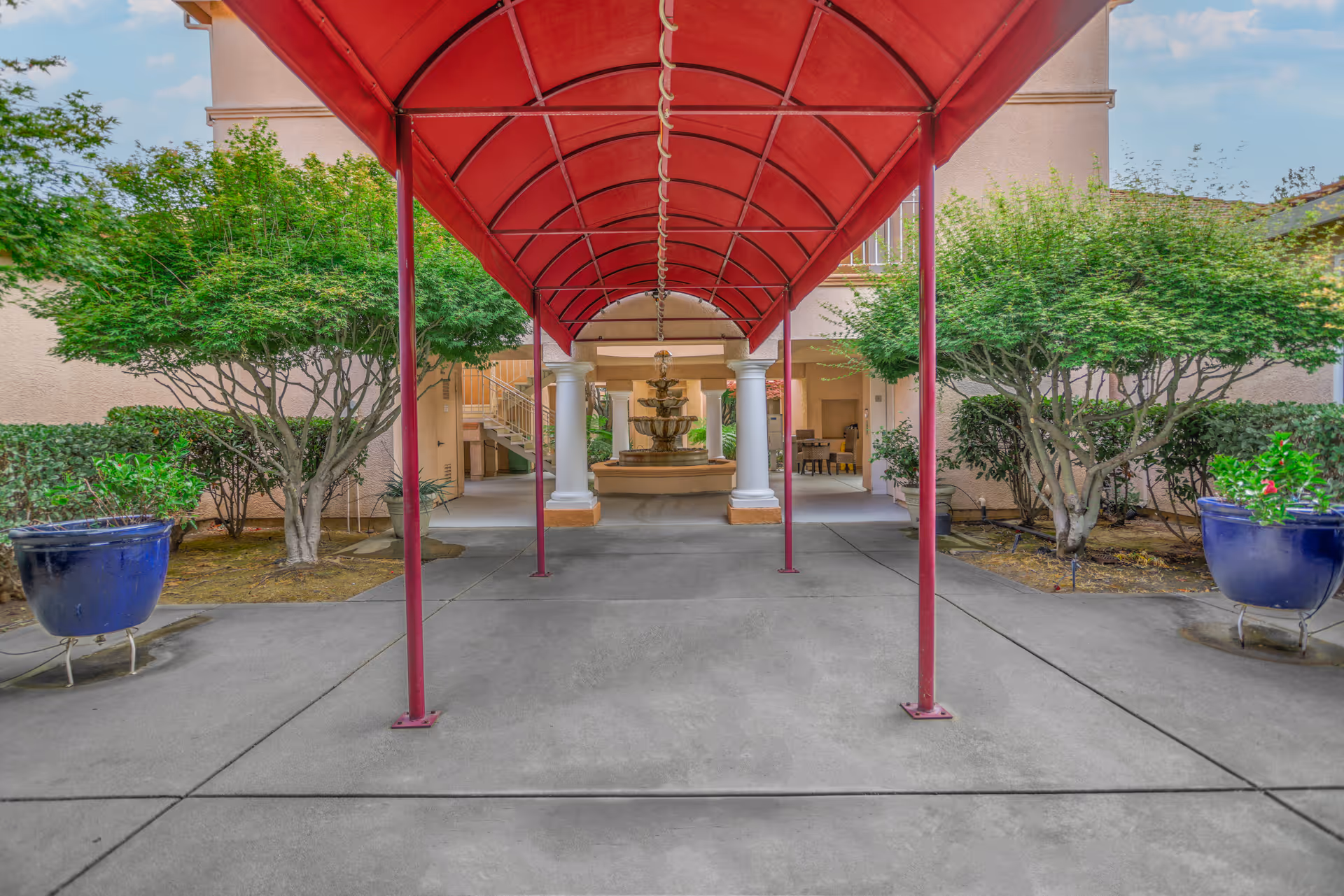 Covered red-canopy walkway leading to a fountain and building entrance framed by columns, potted plants, and trees.