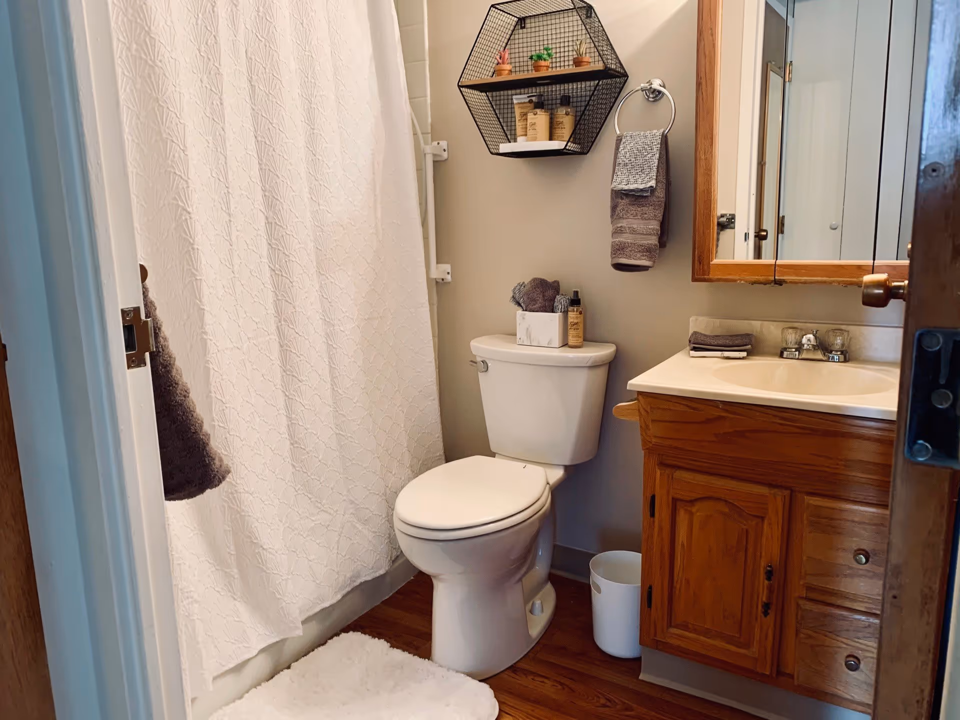 A small bathroom featuring a white toilet, a wooden vanity with a sink, a mirror above the sink, a white shower curtain, and a hexagonal wall shelf holding small plants and toiletries. There are towels hanging on a ring and a hook, and a white bath mat on the wooden floor.