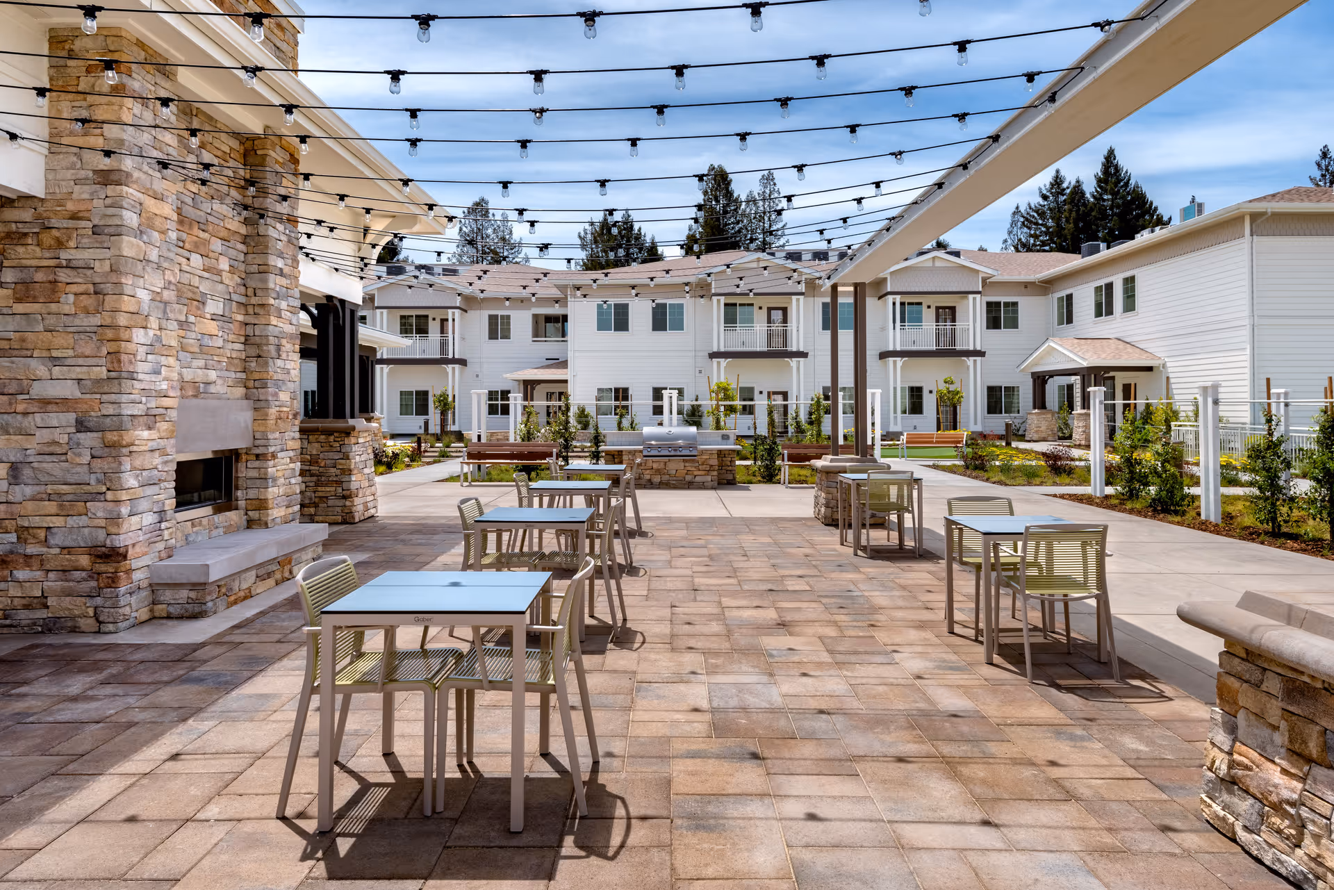 Outdoor patio area at Clearwater at Sonoma Hills featuring several tables with chairs, string lights overhead, a stone fireplace on the left, and a grill in the center background. The patio is surrounded by a two-story white building with balconies and landscaped greenery.