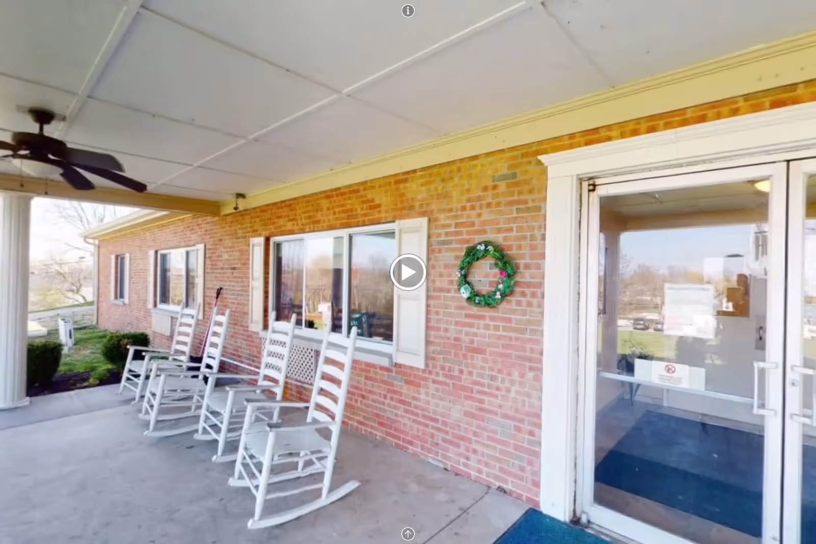 Covered outdoor porch area with a ceiling fan, white rocking chairs lined up against a brick wall, a green wreath hanging on the wall, and glass double doors leading inside a building.
