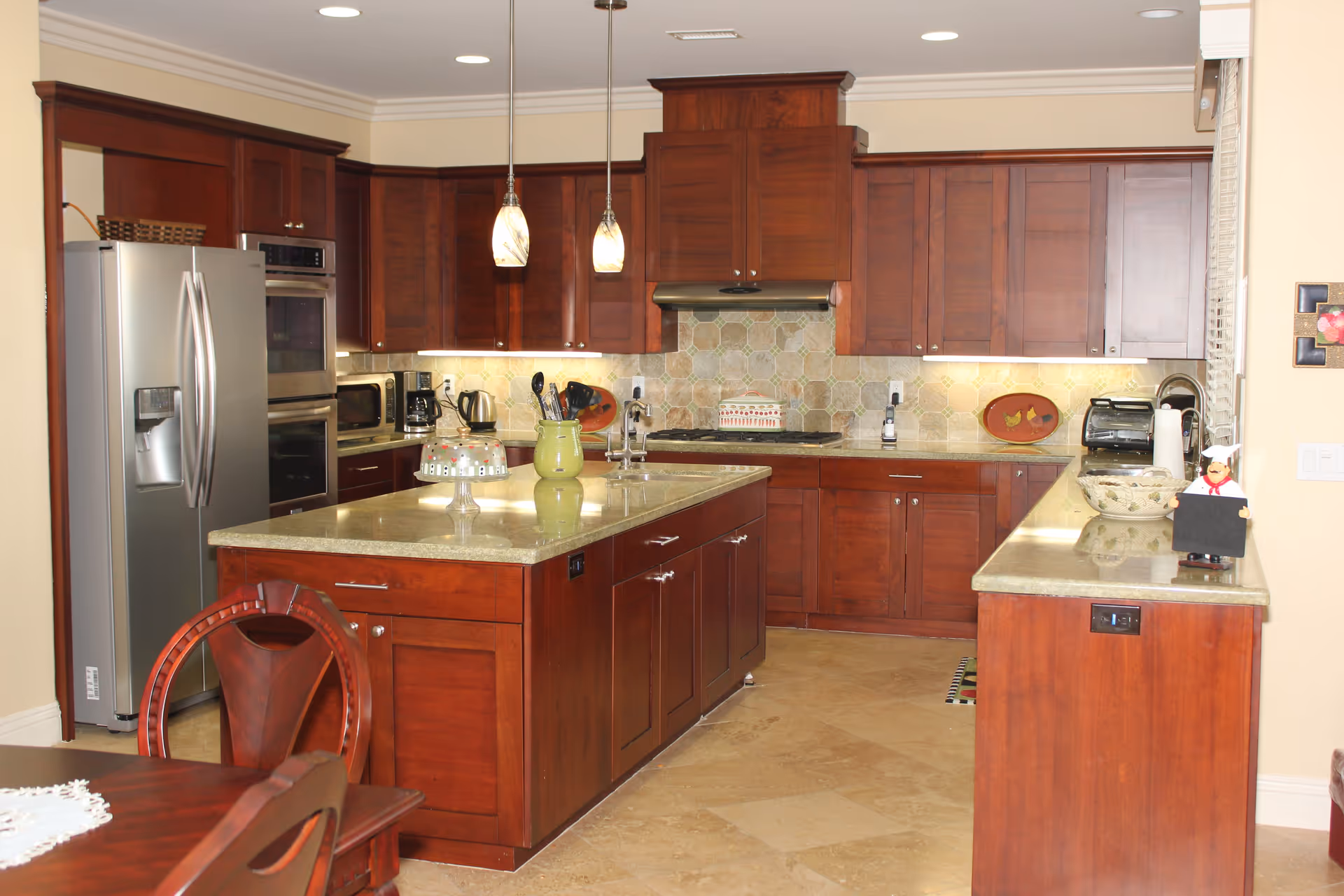 A spacious kitchen with dark wooden cabinets, a large island with a beige countertop, stainless steel refrigerator, double oven, microwave, coffee maker, and various kitchen utensils. Two pendant lights hang above the island, and there is a dining table with chairs partially visible in the foreground.
