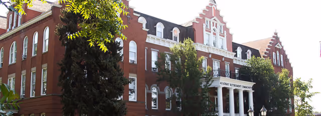 Exterior view of a large, historic red brick building with white columns at the entrance and multiple windows, partially obscured by trees and greenery.