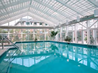 Indoor swimming pool with clear blue water inside a glass-enclosed structure with a high arched ceiling. There are metal handrails leading into the pool, and outside the glass walls, residential buildings and greenery are visible.