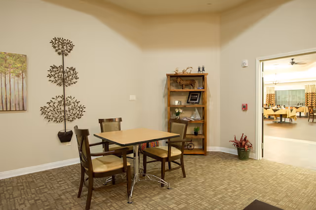 A small seating area with a square wooden table and four chairs with cushioned seats. On the wall to the left is a metal tree wall decoration and a framed painting of trees. To the right is a wooden bookshelf with decorative items including a small deer figurine, a framed photo, and plants. The room has beige walls and carpeted flooring. An open doorway leads to a larger dining area with tables and chairs visible.
