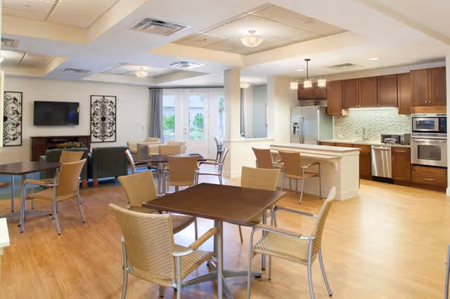 A bright and spacious common area featuring a kitchen with wooden cabinets, stainless steel appliances, and a breakfast bar with stools. The room includes multiple tables and chairs arranged for dining or socializing, with a television mounted on the wall and decorative wall art. Large windows and glass doors allow natural light to fill the space.