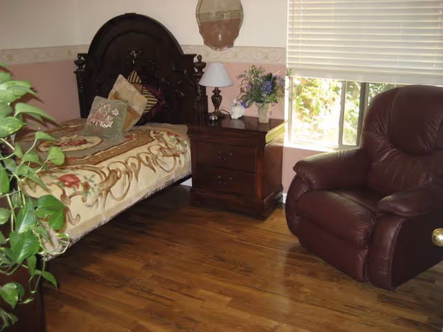 Cozy bedroom with a carved wooden bed, nightstand and lamp, potted plants, a leather recliner, and a window with blinds.