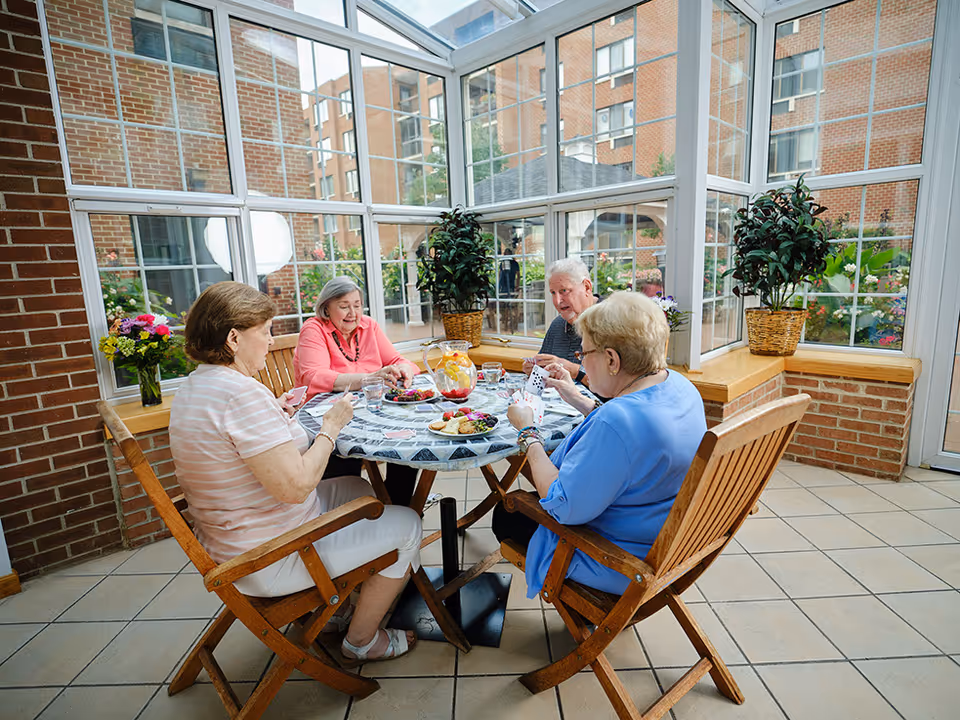 Four elderly people sitting around a round table playing cards in a bright sunroom with large windows and brick walls. The table has a pitcher of lemonade and plates with snacks. There are potted plants and flowers around the room.