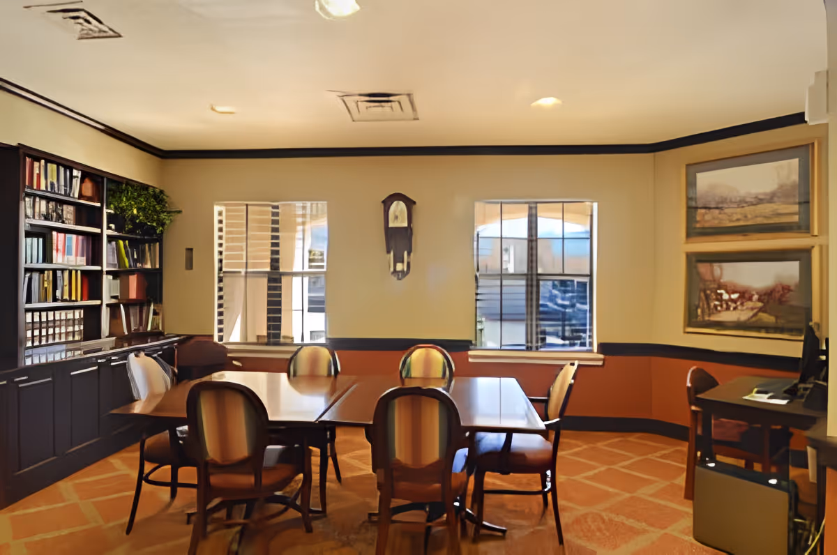 A well-lit meeting or conference room with a large wooden table surrounded by six upholstered chairs. The room has two windows letting in natural light, a wall clock between the windows, built-in dark wood shelves filled with books and decorative items on the left, and two framed landscape paintings on the right wall. The carpet has a geometric pattern in warm tones.