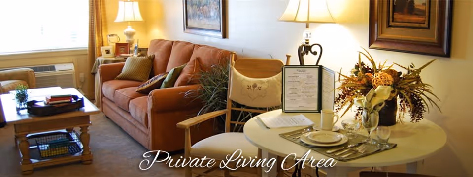 A cozy living area with a brown sofa adorned with cushions, a wooden coffee table with books and a plant, a round dining table set with plates, glasses, and a floral centerpiece, and a lamp providing warm lighting. There are framed paintings on the walls and a window with curtains allowing natural light in.