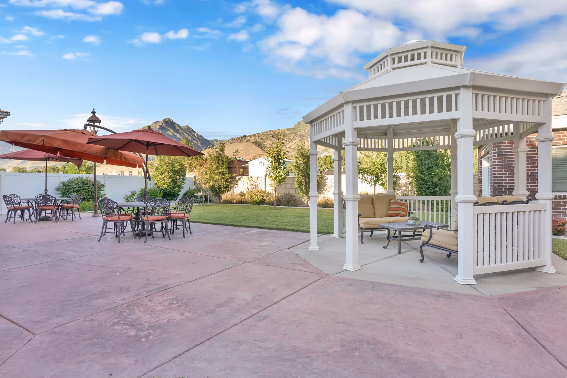 Outdoor patio area with a white gazebo furnished with cushioned seating and a coffee table. Nearby are several metal tables and chairs with red umbrellas providing shade. The background shows a green lawn, trees, a white fence, and mountains under a partly cloudy sky.