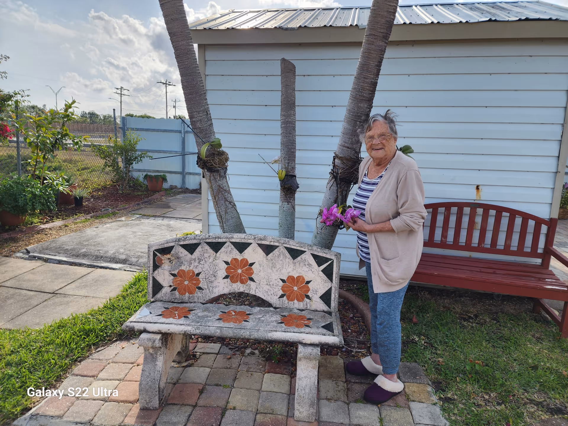An elderly woman standing outdoors next to a decorative stone bench with flower patterns. She is holding purple flowers and smiling. Behind her is a red wooden bench and a white building with three palm tree trunks in front. The area is paved with bricks and surrounded by grass and plants.