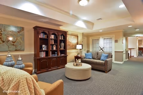 Comfortable communal living room with a sofa, armchairs, round ottoman, bookshelf, and soft overhead lighting.