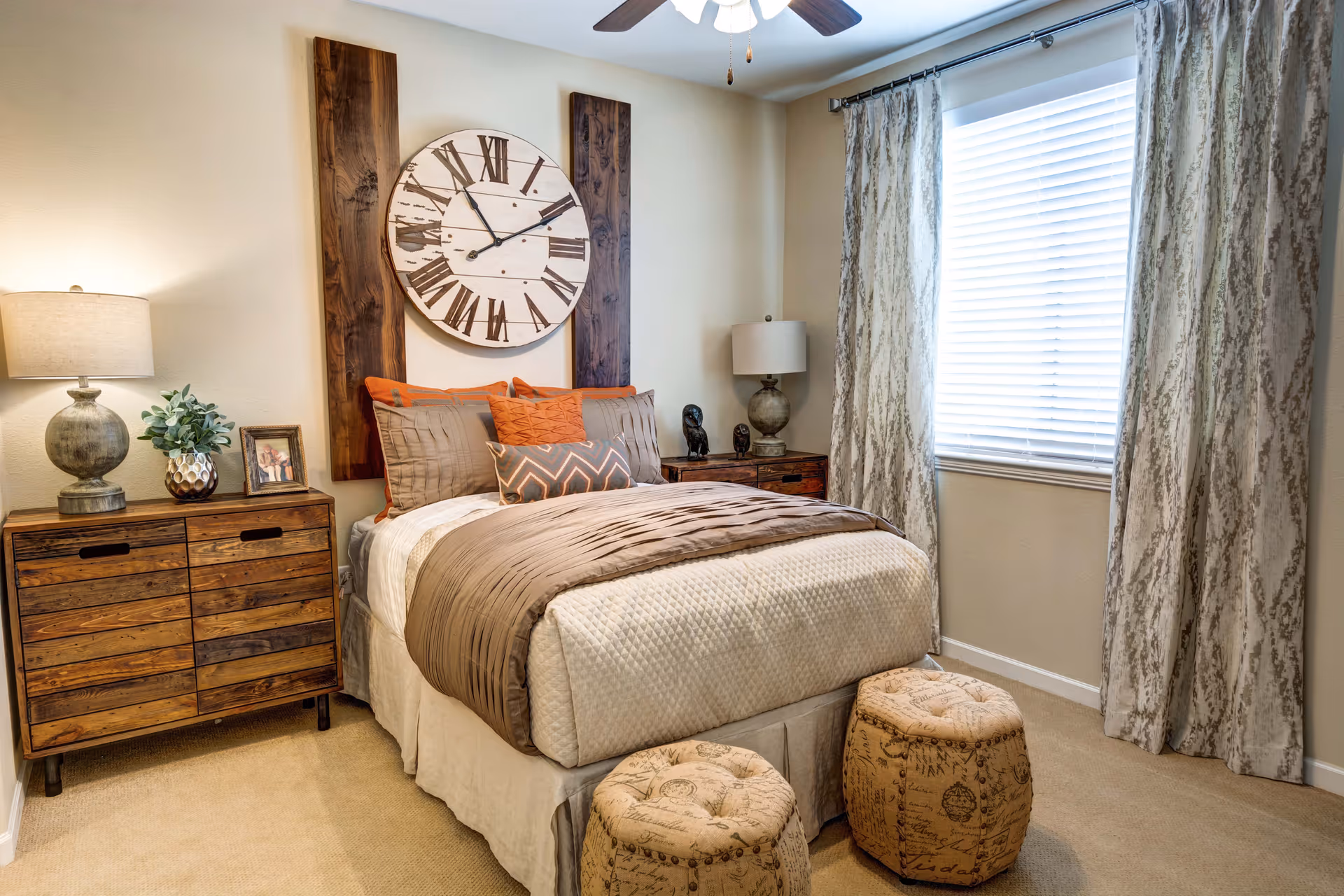 A cozy bedroom with a neatly made bed featuring beige and brown bedding and orange accent pillows. Above the bed is a large round clock with Roman numerals mounted on two vertical wooden panels. On either side of the bed are wooden nightstands with lamps, a small plant, and decorative items. A window with patterned curtains allows natural light into the room. Two round, patterned ottomans are placed at the foot of the bed.