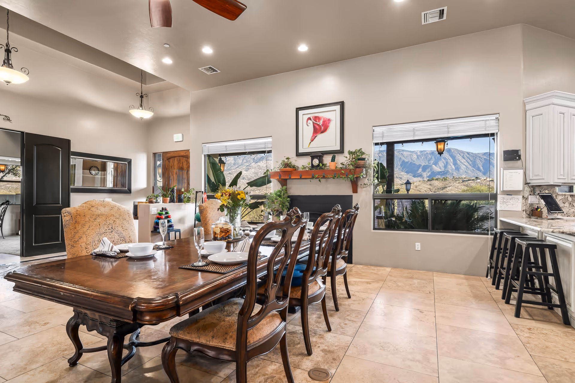 A spacious dining room with a large wooden dining table set for six, featuring elegant chairs and tableware. The room has beige tiled floors, light-colored walls, and large windows offering a scenic view of mountains outside. There is a fireplace with a wooden mantel decorated with plants and a framed flower artwork above it. To the right, a kitchen counter with bar stools is visible, and the room is well-lit with ceiling lights and hanging lamps.