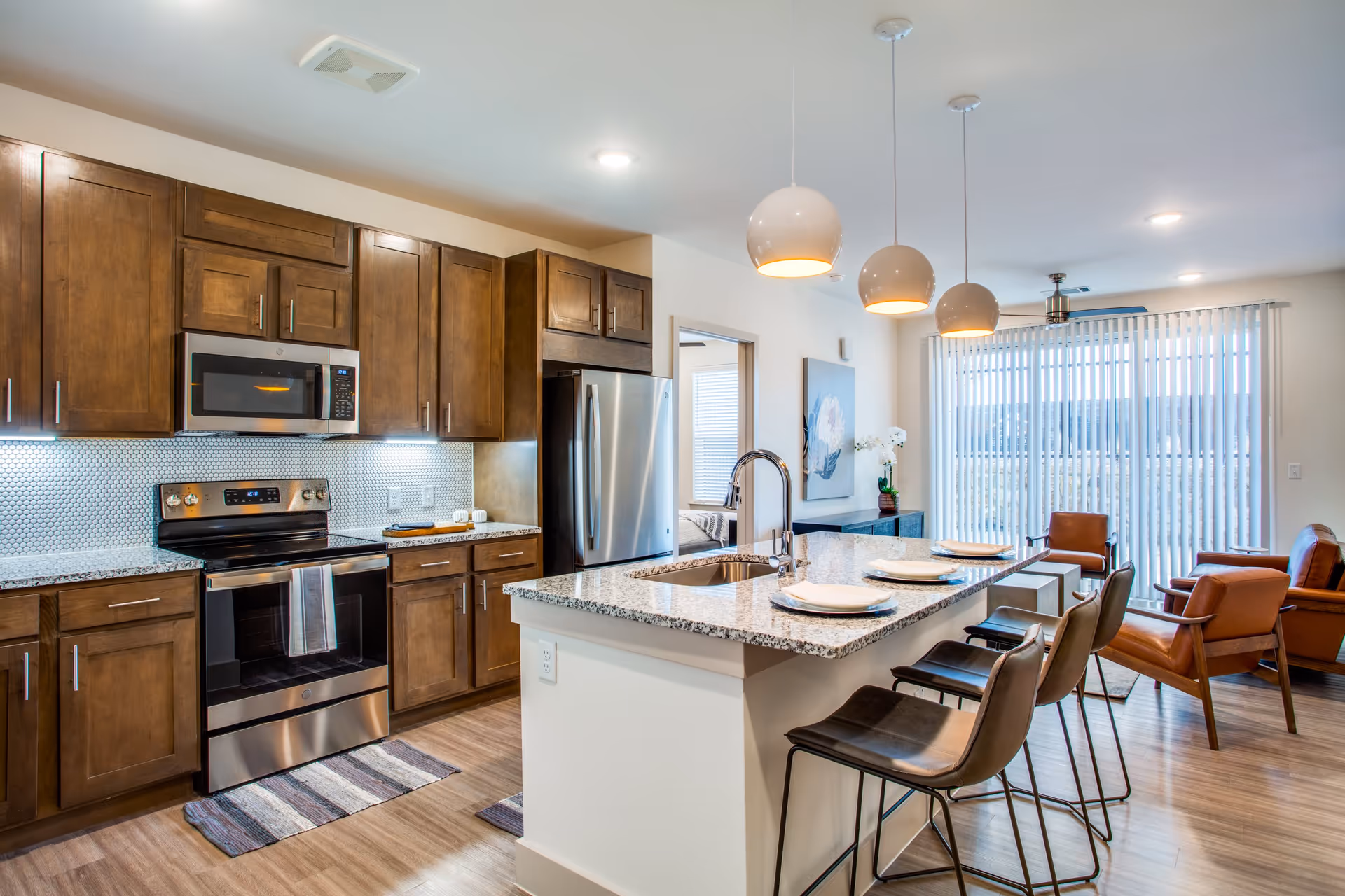Modern kitchen and living area with wooden cabinets, stainless steel appliances including a microwave, stove, and refrigerator. A granite countertop island with a sink and four bar stools is set with plates and napkins. The living area has leather chairs and large vertical blinds covering a window or sliding door.