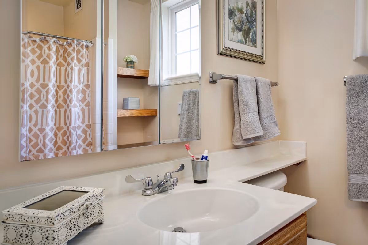 A clean bathroom with a white sink and countertop, a silver faucet, a toothbrush and toothpaste in a metal cup, two gray towels hanging on a towel rack, a decorative box on the counter, a window with white curtains, a framed floral picture on the wall, and a shower with a patterned curtain.
