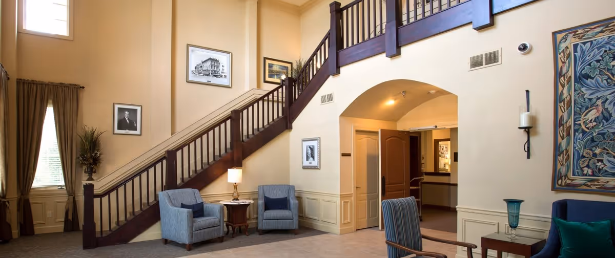 Interior view of a senior living facility lobby with a staircase featuring dark wooden railings. There are two blue armchairs with a small wooden table and lamp between them, a striped chair, framed pictures on the walls, and a large decorative tapestry. The walls are painted beige and there is a window with brown curtains letting in natural light.