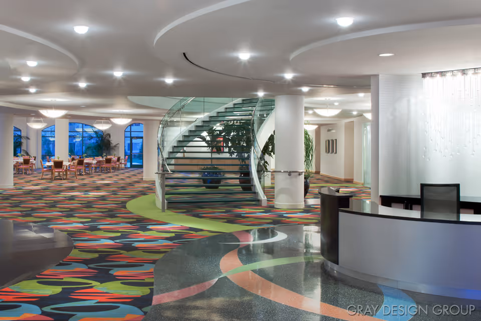 Bright modern lobby featuring a curved reception desk, a glass spiral staircase, and a colorful patterned carpet leading to a dining area.