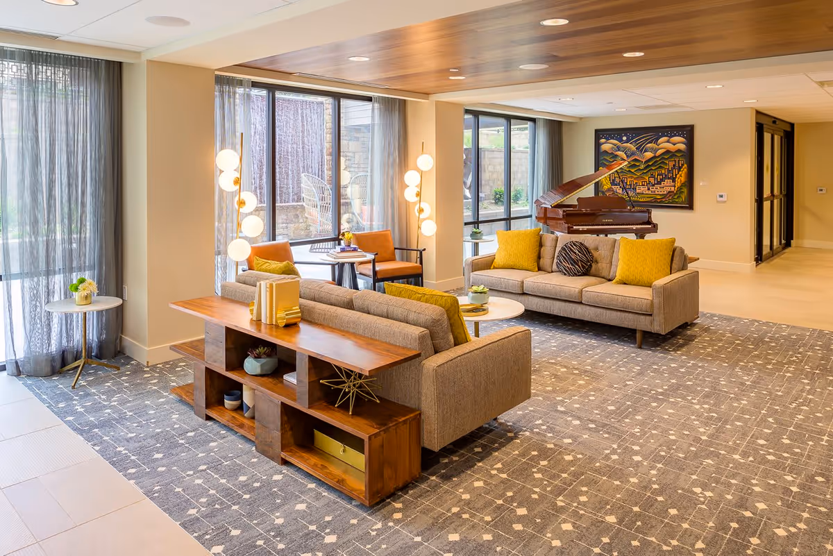 A bright and spacious senior living common area featuring two beige sofas with yellow and patterned cushions, a wooden console table with decorative items, two orange armchairs with a small round table between them, modern floor lamps with globe lights, large windows with sheer curtains, a grand piano, and a colorful painting on the wall.