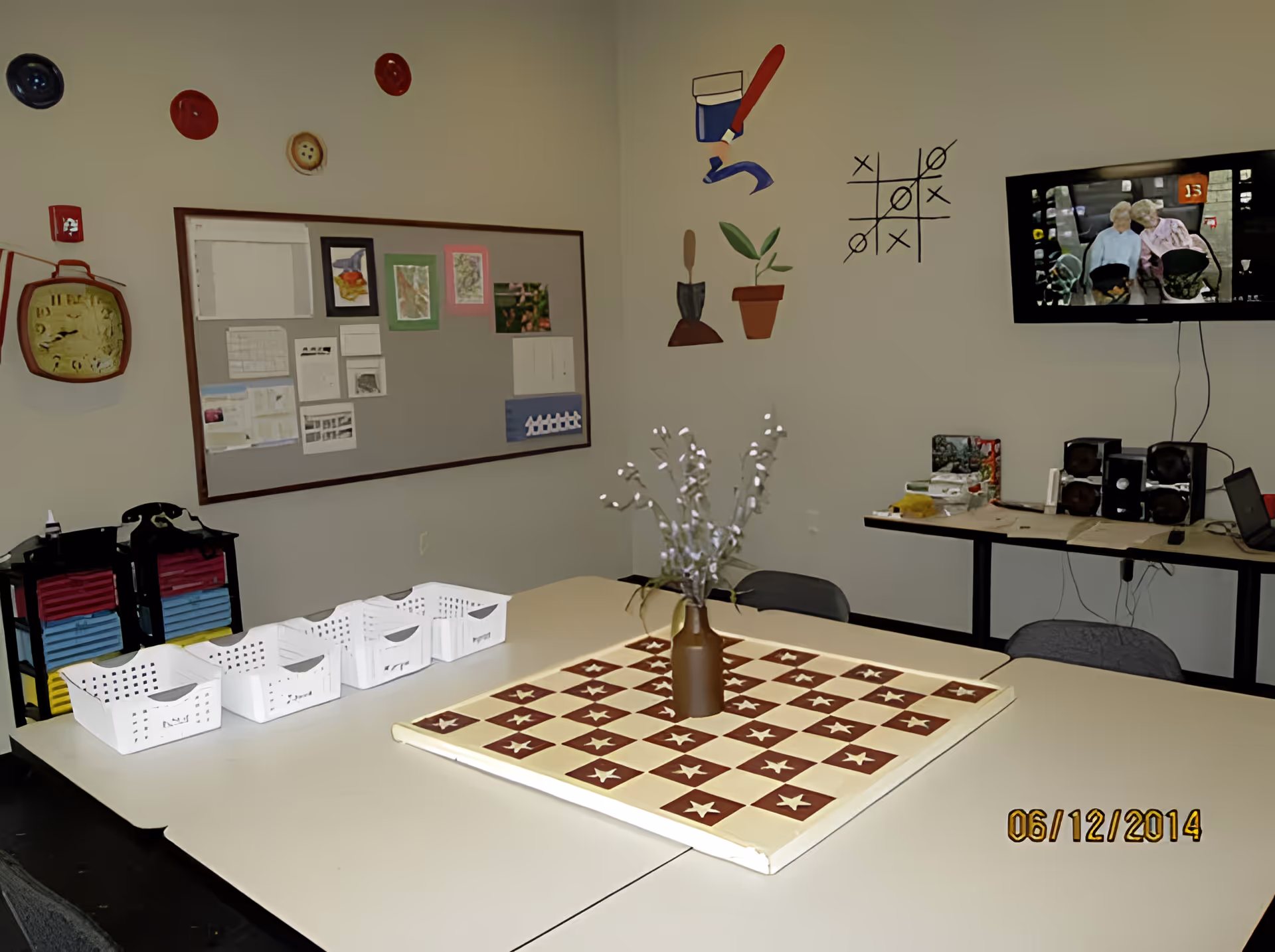 A room with a large table in the center featuring a checkerboard pattern with stars and a vase with white flowers. On the left wall, there is a bulletin board with various papers and artwork pinned to it, along with decorative buttons and a clock. The right wall has painted images of a paint tube, a trowel, a potted plant, and a tic-tac-toe game. A TV mounted on the wall displays an image of two elderly people. There are several chairs around the table and a side table with speakers and other items.