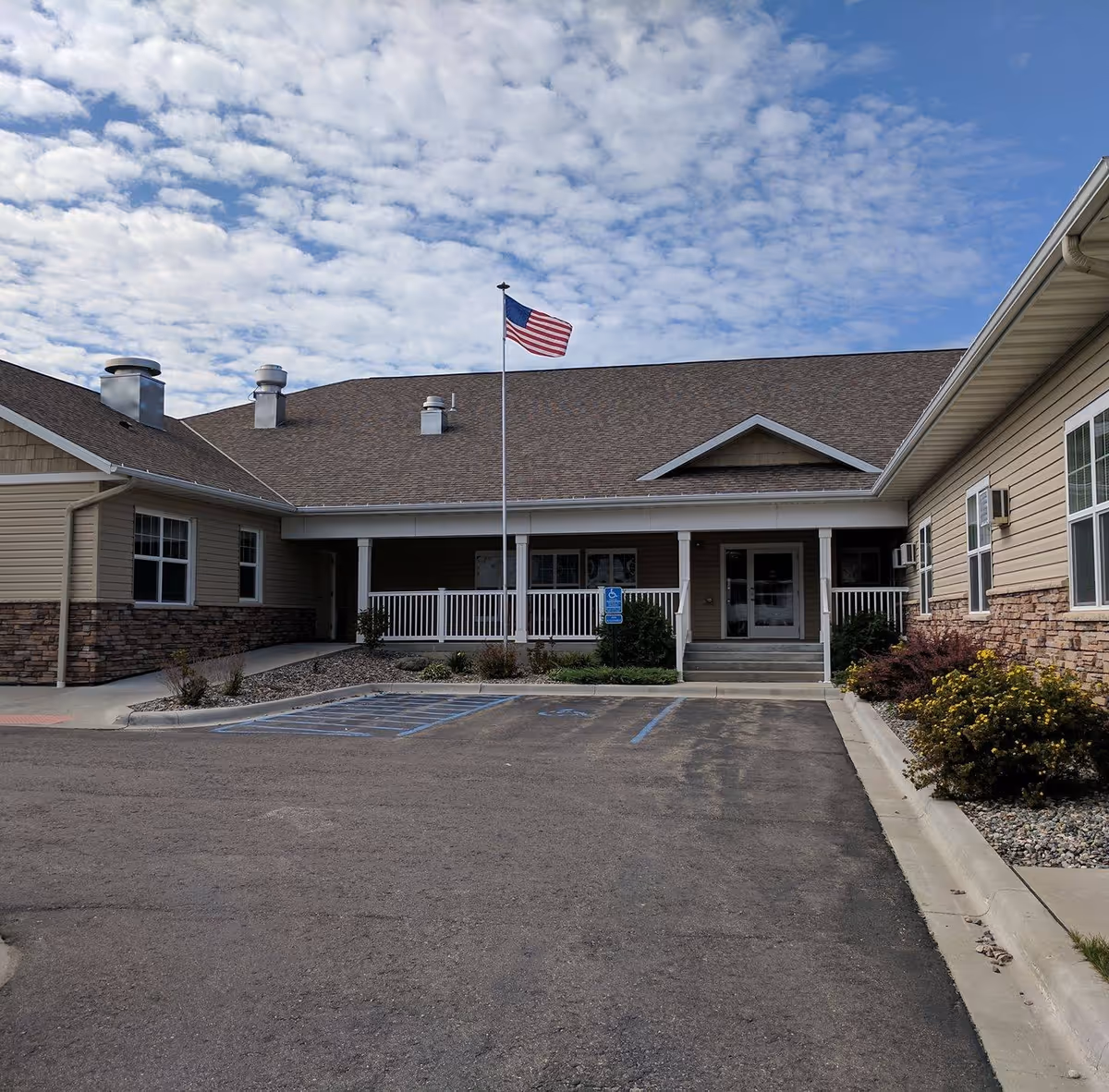 Exterior view of a single-story senior care facility building with beige siding and stone accents. There is a covered entrance with white railings and steps leading up to double doors. An American flag is flying on a flagpole in front of the entrance. The sky is partly cloudy and there are some bushes and landscaping around the building. The parking lot in front has marked handicap parking spaces.