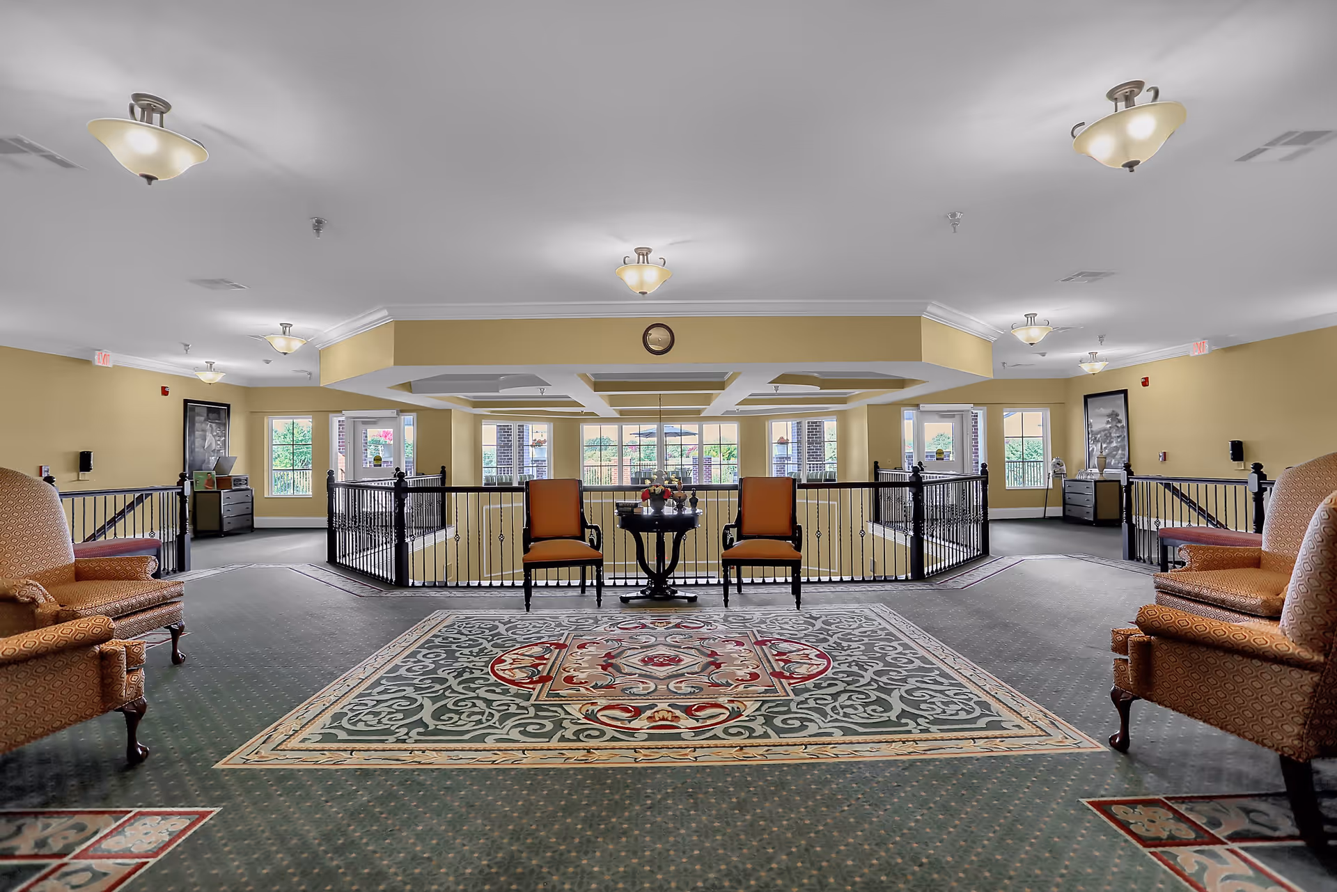 A spacious and well-lit common area in a senior living facility featuring a large decorative carpet, two orange upholstered chairs with a small round table between them, and additional armchairs on either side. The area overlooks a lower level through black metal railings, with large windows allowing natural light to fill the space. The walls are painted a soft yellow, and ceiling lights provide additional illumination.