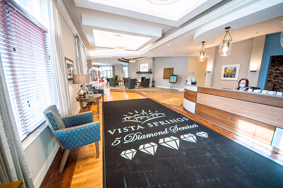 Reception area of Vista Springs Ravinia Estate featuring a wooden front desk with a receptionist wearing a mask, a black floor mat with the Vista Springs 5 Diamond Services logo, blue patterned chairs, a lamp on a small table, large windows with blinds, and a seating area with chairs and a fireplace in the background.